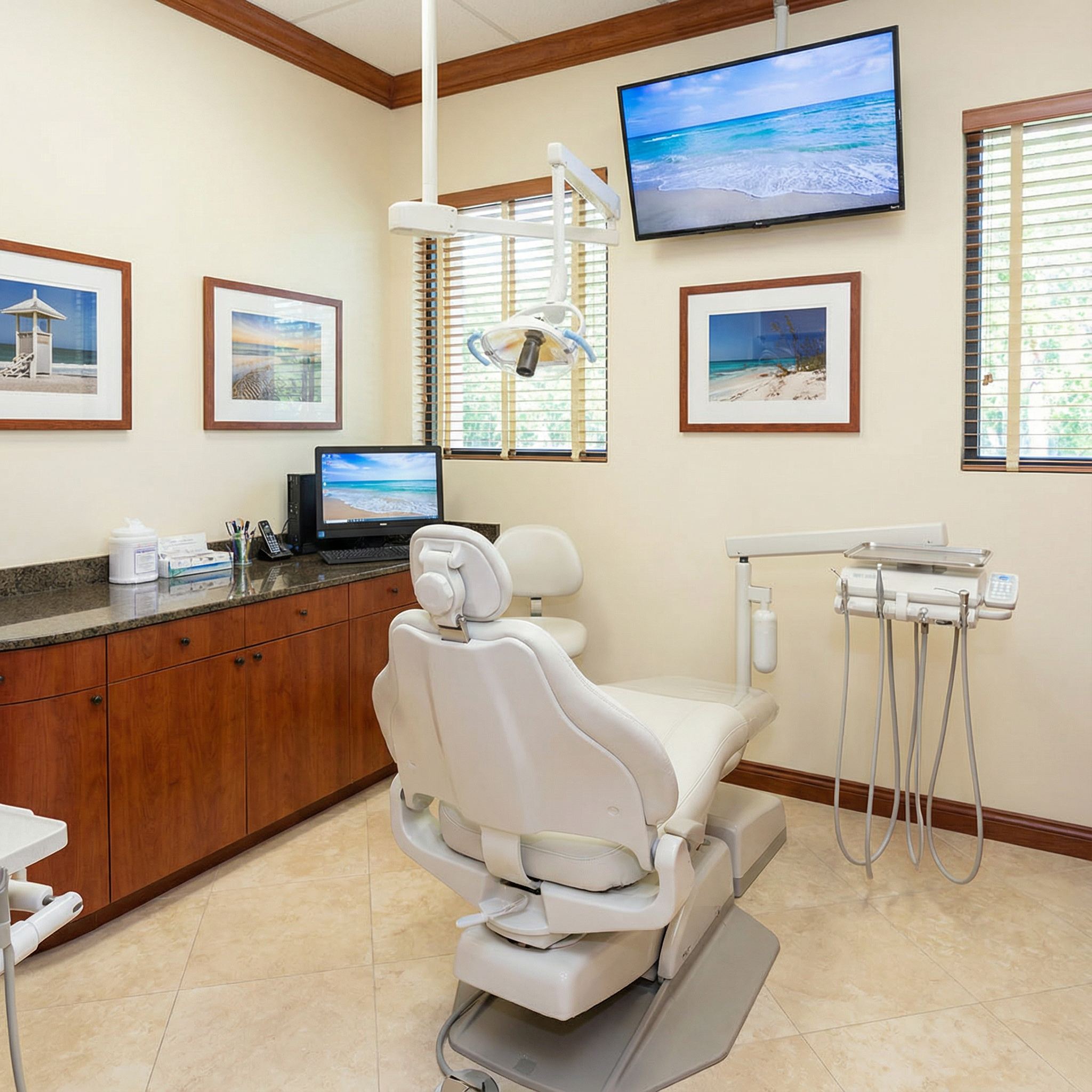 Treatment room with white dental chair, beach-themed artwork, TV monitor, and cherry wood cabinets